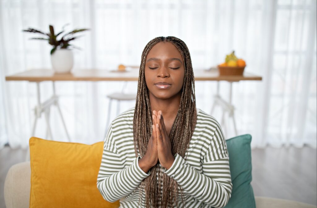 Peaceful black woman sitting on couch at home, praying